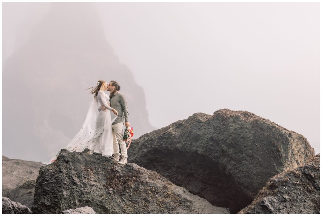 A couple kissing on a a rock on at black sand beach with her wedding dress flowing in the breeze.
