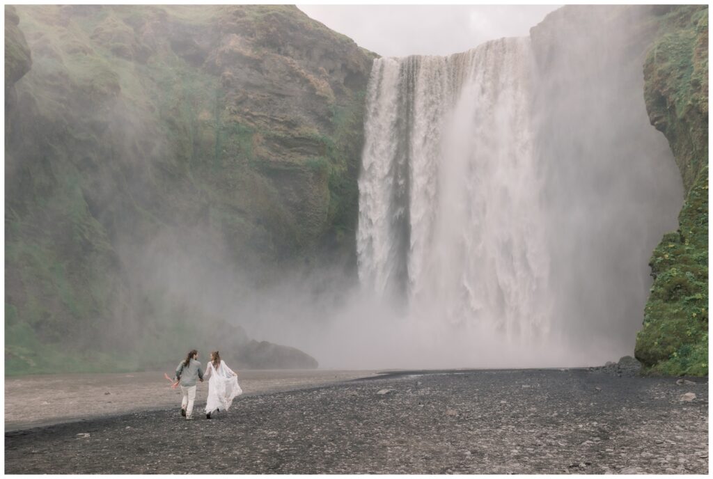 Man and woman running toward a huge waterfall on their elopement in Iceland.
