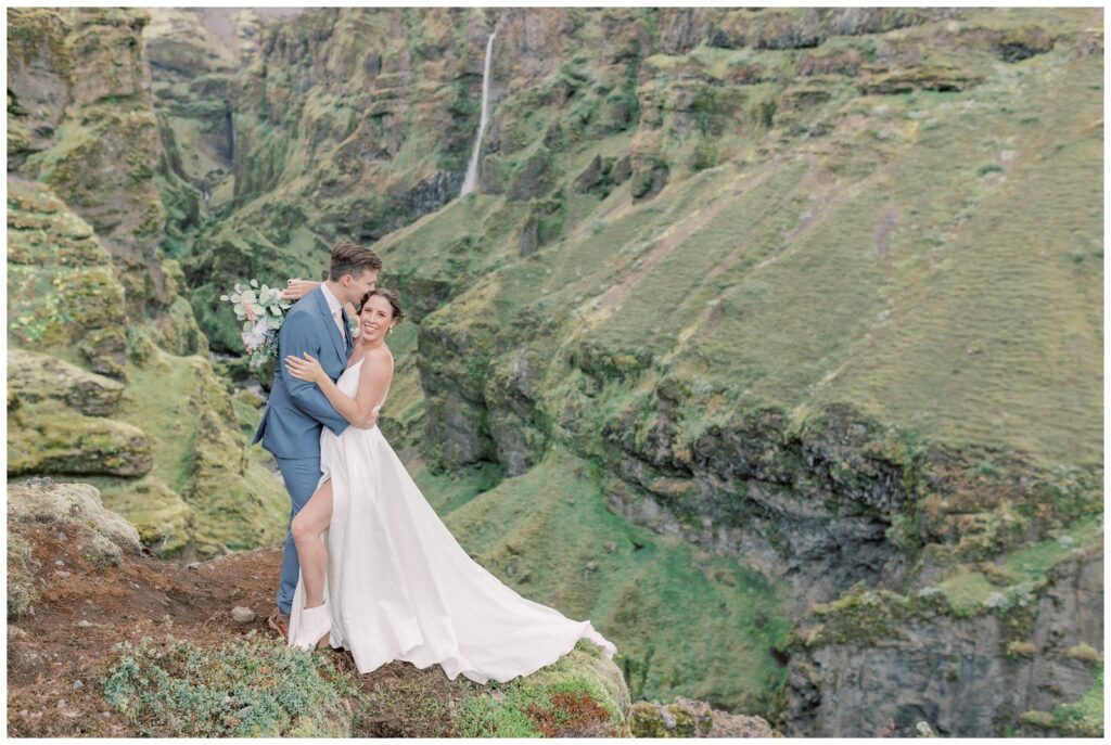 During an Iceland elopement, this couple hiked to a hidden canyon. The groom is kissing his bride's head as they take in the view of the waterfall surrounded by mountains. 