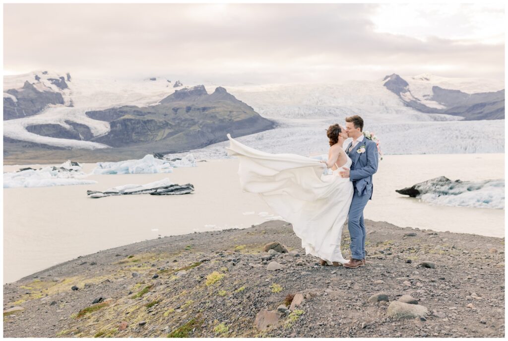 A couple kissing on their elopement with glaciers in the background. The bride's dress if flowing in the wind during this couple's Iceland Elopement.