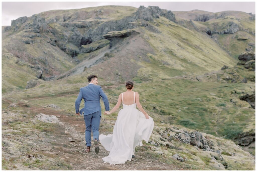 Bride and groom holding hands while running up a mountain in their wedding attire during their elopement in Iceland.
