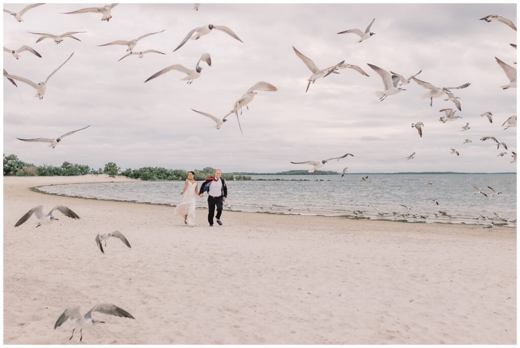 Bride and groom holding hands and running along the shore line at a beach in Crystal River, surrounded by seagulls. Fort Island Gulf Beach is one of the best places to elope in Florida.