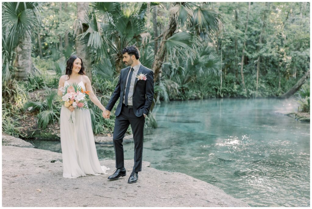 Couple on their wedding day holding hands and smiling at each other at Rock Springs in Kelly Park, one of the best places to elope in Florida. The Springs are behind them with mist floating over the water at sunrise during their Florida elopement.