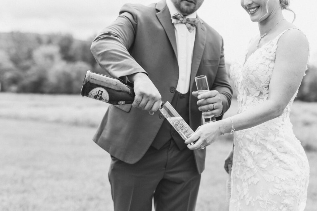 Groom pouring champagne in Bride's glass in a field as one if their activities during their Lake Placid Elopement.
